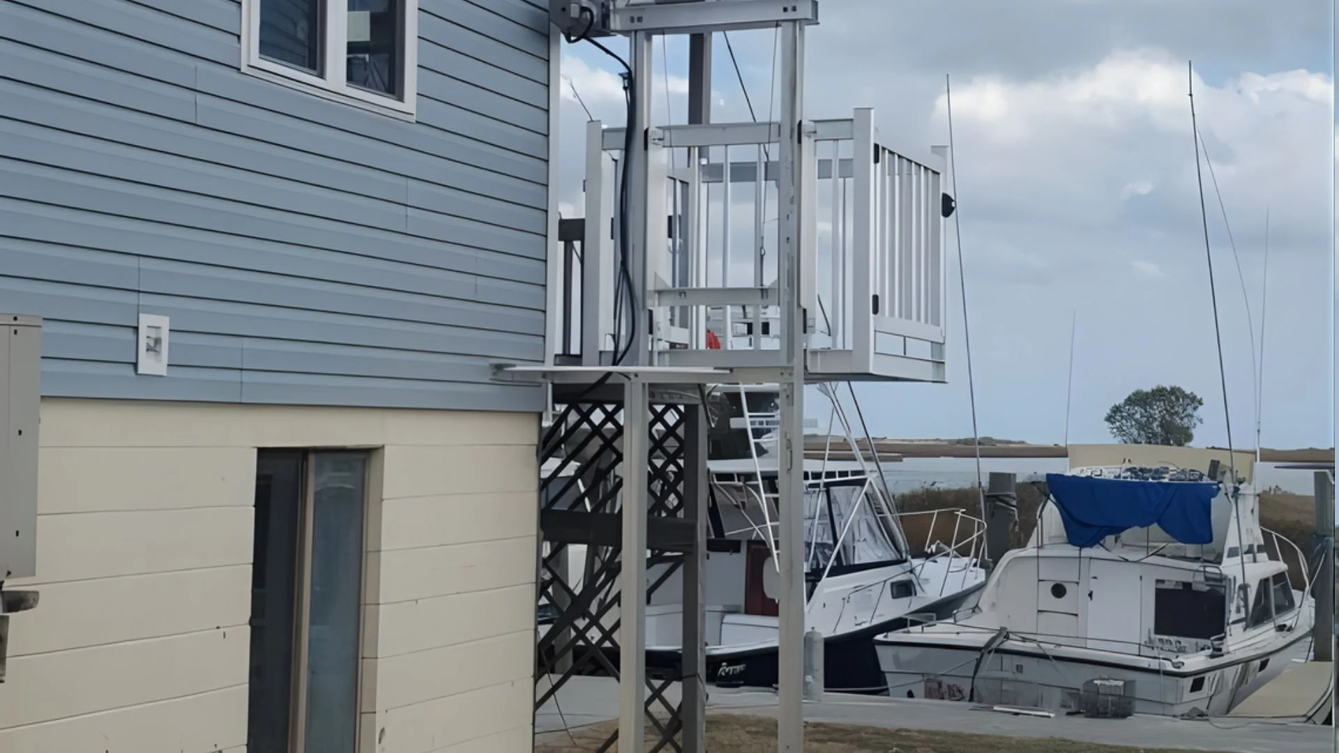Outdoor home elevator next to a blue house by a marina, boats docked nearby under a cloudy sky.