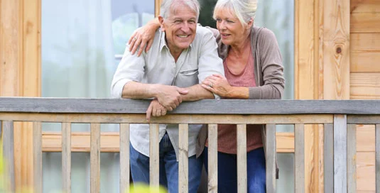 Elderly couple smiling on wooden porch, leaning on railing, with a cozy wooden house in the background.