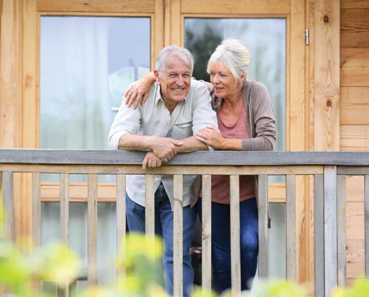 Elderly couple smiling on wooden porch, leaning on railing, with a cozy wooden house in the background.