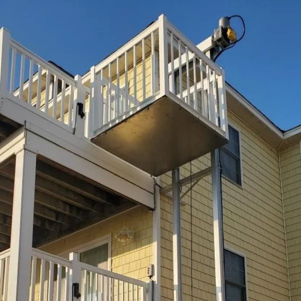 Outdoor residential platform with railing, used as a home elevator on a two-story yellow house, under a clear blue sky.