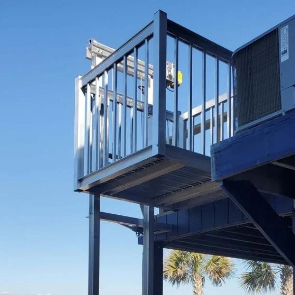 Outdoor home elevator platform with metal railings, partially shaded, against a clear blue sky and a glimpse of palm trees.
