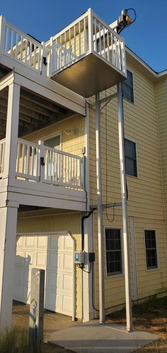 Exterior yellow house with a home elevator leading to an upper balcony. Nearby, a garage door and windows are visible in the sunlit scene.