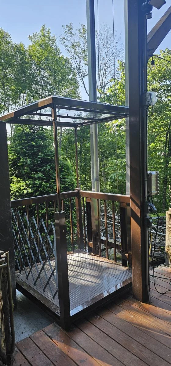 Outdoor wooden platform with a railing, surrounded by trees; resembles a home elevator setup.