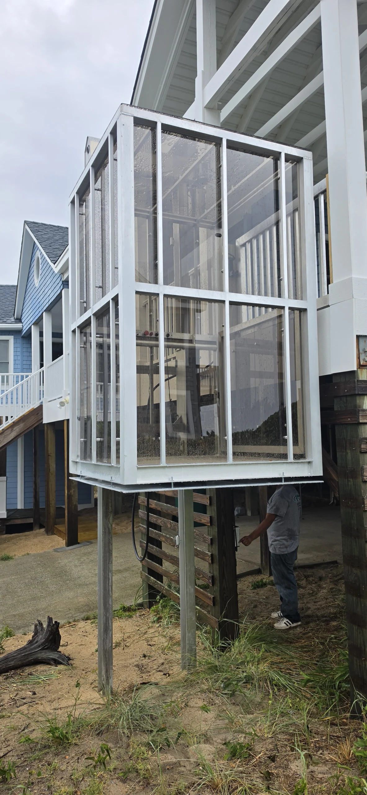 Glass-enclosed home elevator connected to a house on stilts, with a person standing below working on it.