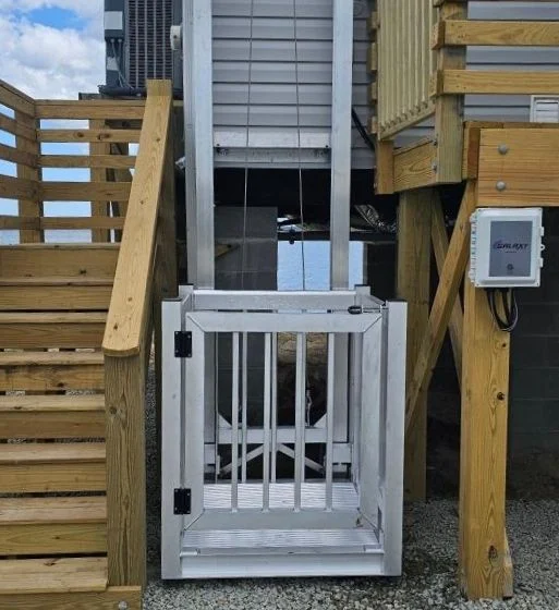 Outdoor home elevator beside wooden stairs, featuring a gated platform and control box on a stilted structure.