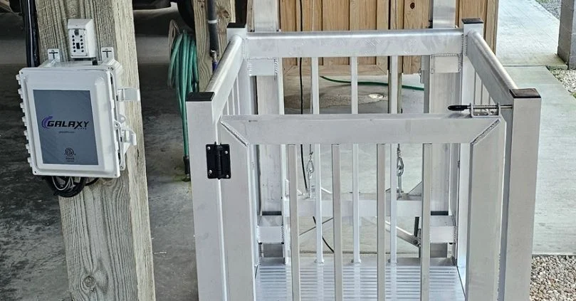Outdoor home elevator with gate open, next to a control box mounted on a wooden post, on a concrete and gravel surface.