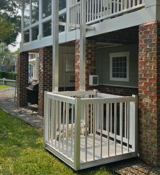Outdoor home elevator with white railings, located under a raised deck with brick pillars, providing accessibility to the upper level.