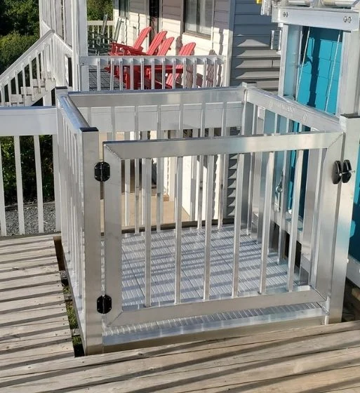 Aluminum home elevator on wooden deck, featuring a gated enclosure, adjacent to stairs and red chairs.