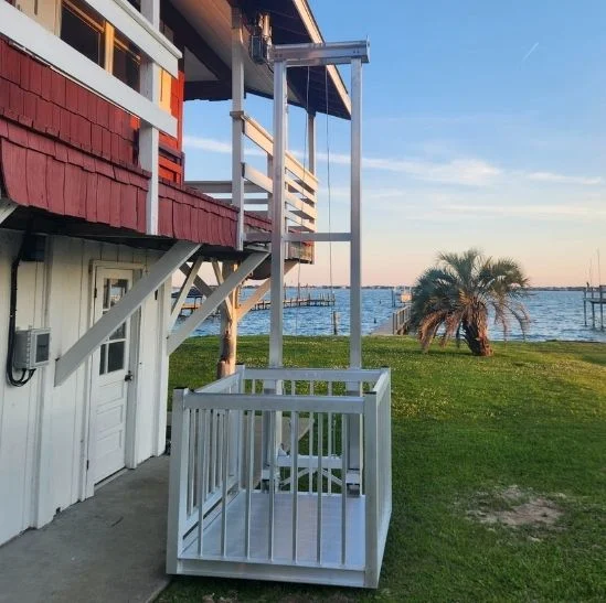 Outdoor home elevator beside a two-story building, overlooking a grassy area and waterfront with a palm tree near the shore.