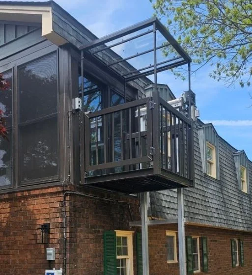 Outdoor home elevator attached to a rustic two-story house, featuring a metal frame and surrounded by greenery.