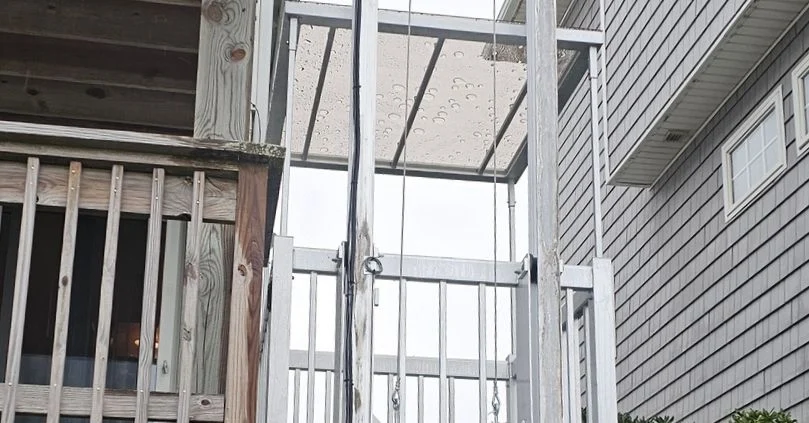 Outdoor home elevator with metal frame, adjacent to a wooden balcony. Transparent roof panel visible above the elevator.