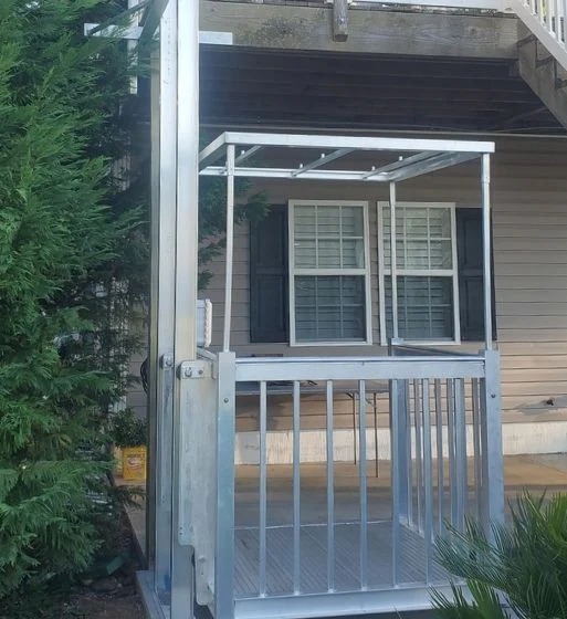 Outdoor home elevator with a metal frame next to a house, enhancing accessibility for the porch area, surrounded by greenery.