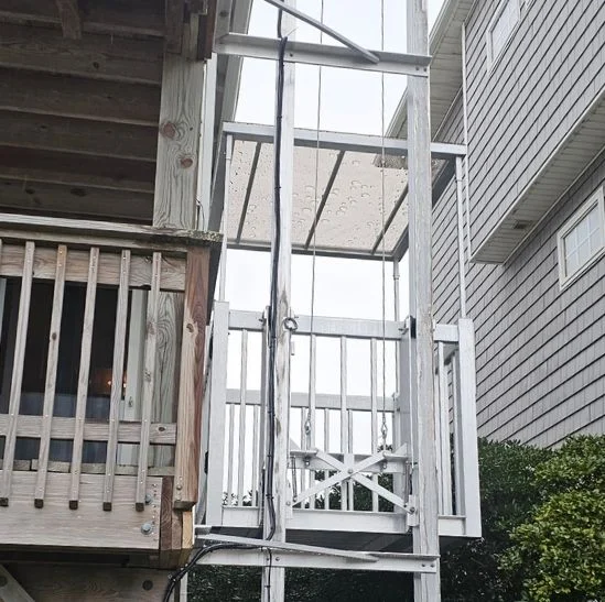 Outdoor home elevator with a white metal frame beside a wooden deck, installed on a multi-story house.