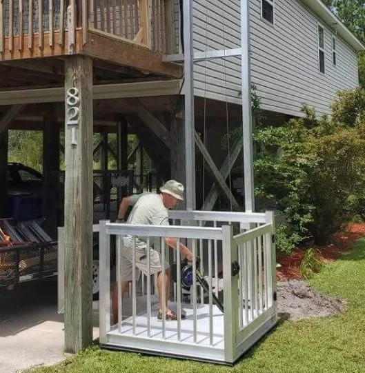 Man in hat helps dog onto outdoor platform-style home elevator under raised house, surrounded by greenery.