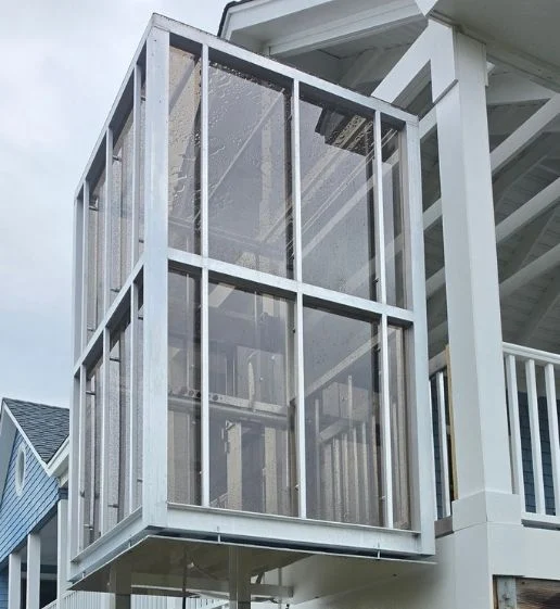 Modern glass-walled home elevator attached to an exterior porch, showing rainy weather marks on the surface.