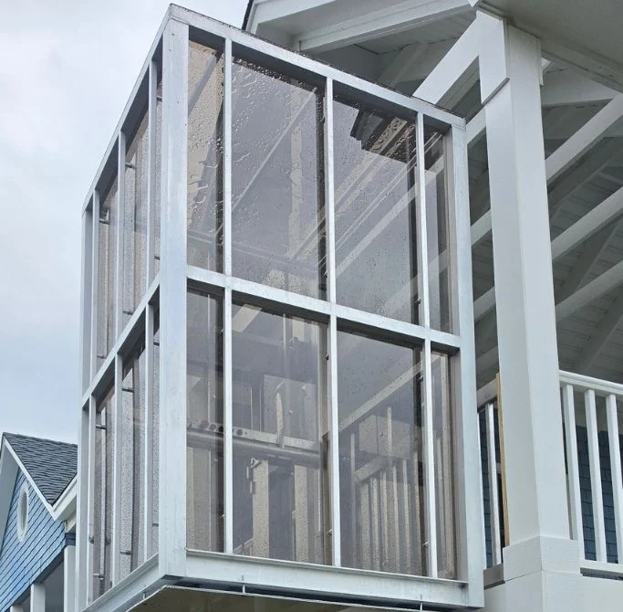 Modern glass-panel home elevator attached to a white house exterior under a cloudy sky, showcasing accessibility features.