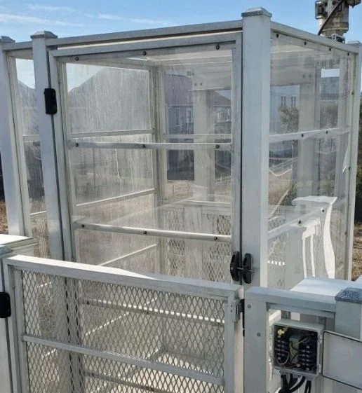 Transparent outdoor home elevator with metal framing, showing control panel and open sides, situated in a residential area.