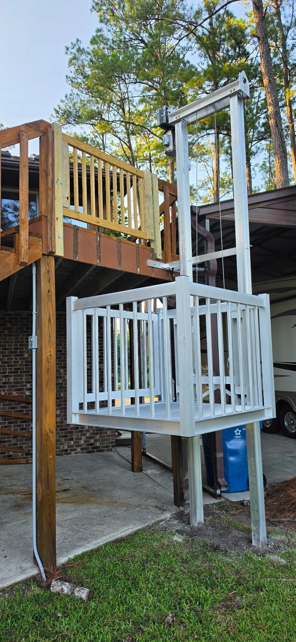 Outdoor home elevator with white railing beneath a wooden deck, surrounded by trees and grass.