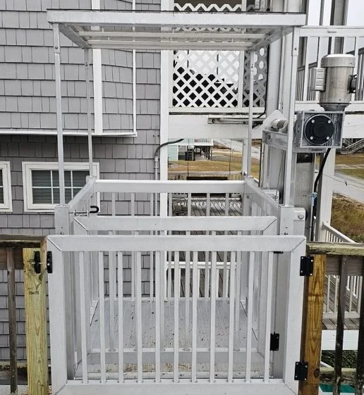 White outdoor home elevator with gated platform next to a grey shingled house, featuring wooden guardrails and a motorized mechanism.