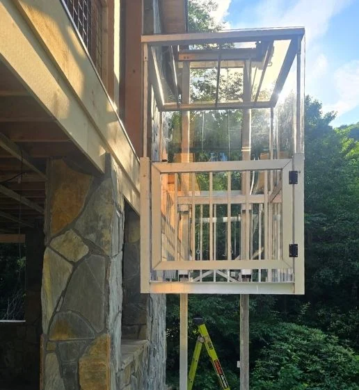 Sunlit construction of a wooden frame structure resembling home elevators attached to a stone wall, with a ladder visible in the background.